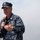 A U.S. Navy servicemember in Navy Working Uniform (NWU Type I) digital camouflage is speaking into a handheld microphone, likely addressing an audience at an outdoor event. The individual wears visible rank/rating insignia on their cap and chest, along with a qualification badge consistent with submarine warfare. The background is overcast and hazy, suggesting an outdoor setting near water or an open area.