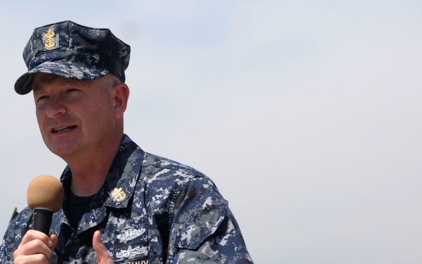 A U.S. Navy servicemember in Navy Working Uniform (NWU Type I) digital camouflage is speaking into a handheld microphone, likely addressing an audience at an outdoor event. The individual wears visible rank/rating insignia on their cap and chest, along with a qualification badge consistent with submarine warfare. The background is overcast and hazy, suggesting an outdoor setting near water or an open area.
