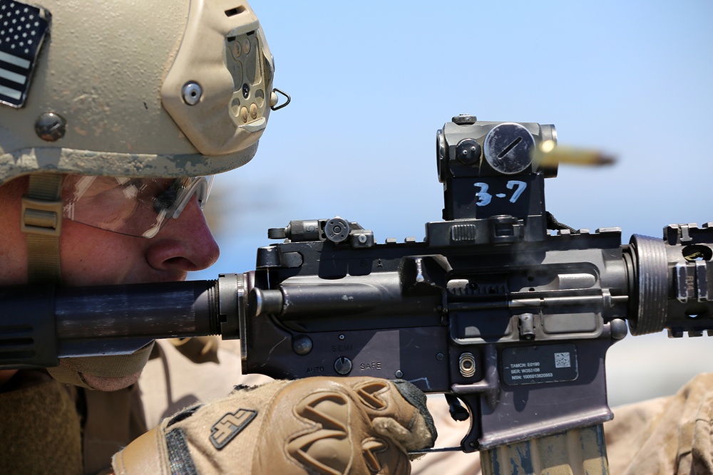 A U.S. military service member is actively firing an M4 carbine rifle, captured mid-shot with a spent brass casing visibly ejecting from the weapon. The soldier is wearing standard U.S. Army combat gear including a tan MICH/ACH helmet with an American flag patch, ballistic eyewear, and tan tactical gloves. The rifle is marked '3-7' on the optic mount, consistent with unit identification markings.