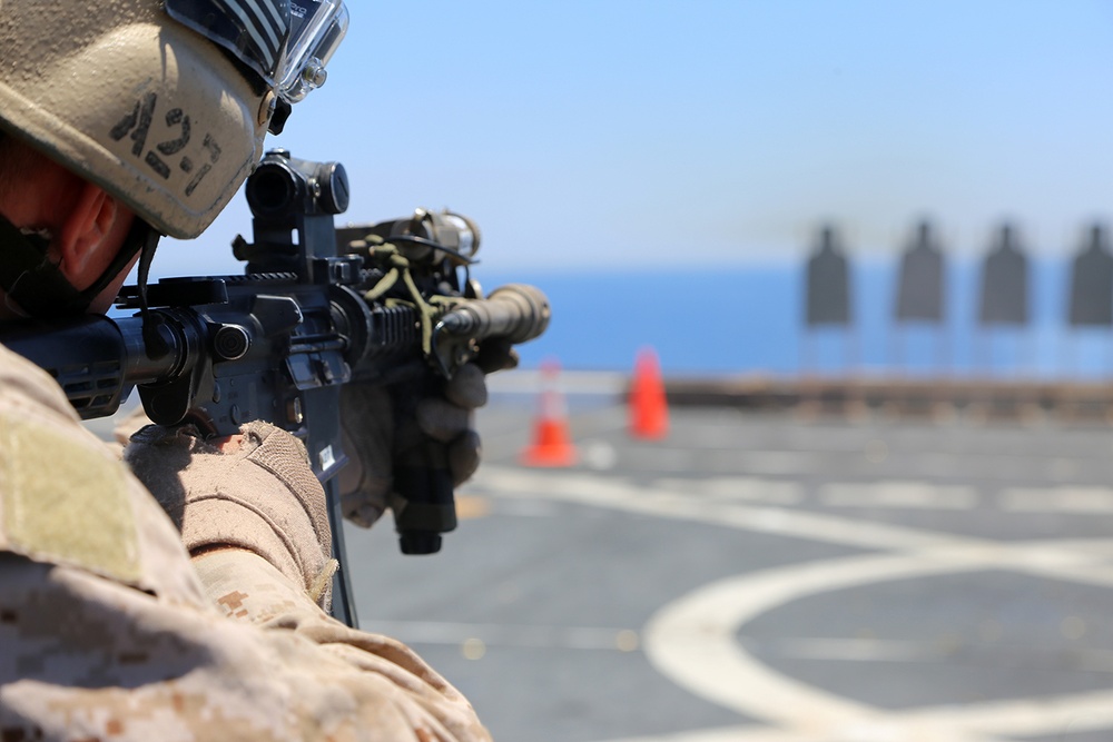 A military service member in desert camouflage uniform and helmet is aiming an assault rifle on what appears to be a ship's flight deck or helipad, indicated by the circular markings on the deck surface. Several human-silhouette shooting targets are visible downrange, along with orange traffic cones marking the range boundaries. The setting suggests a live-fire marksmanship training exercise conducted at sea.