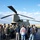 A group of civilians is gathered on a tarmac around a military CH-47 Chinook tandem-rotor helicopter, appearing to receive a briefing or tour from uniformed personnel. The setting appears to be an airfield or military base, with flat grassland visible in the background under a partly cloudy sky. This appears to be a public outreach, media day, or educational event.