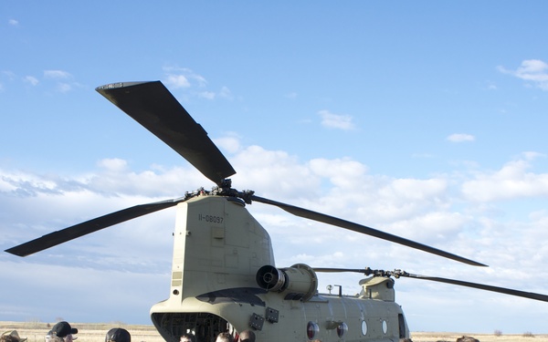 A group of civilians is gathered on a tarmac around a military CH-47 Chinook tandem-rotor helicopter, appearing to receive a briefing or tour from uniformed personnel. The setting appears to be an airfield or military base, with flat grassland visible in the background under a partly cloudy sky. This appears to be a public outreach, media day, or educational event.