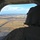 A military crew member wearing a flight helmet and combat uniform is seated at the open rear ramp of a large military helicopter or transport aircraft, looking out over a flat, agricultural landscape below. A runway or airstrip is visible on the ground, suggesting the aircraft is either approaching or departing from a small airfield. The terrain appears to be open plains or farmland under partly cloudy skies.