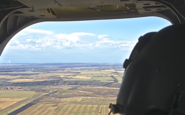 A military crew member wearing a flight helmet and combat uniform is seated at the open rear ramp of a large military helicopter or transport aircraft, looking out over a flat, agricultural landscape below. A runway or airstrip is visible on the ground, suggesting the aircraft is either approaching or departing from a small airfield. The terrain appears to be open plains or farmland under partly cloudy skies.
