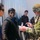 A Canadian Armed Forces soldier in full combat uniform and gear is checking documents, likely a passport, presented by two civilians. The interaction appears to be a checkpoint or processing scenario, possibly a military exercise or humanitarian operation. A third civilian and another individual in tactical gear are visible in the background.