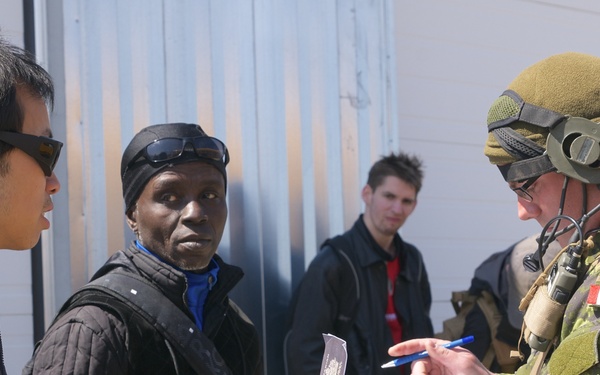 A Canadian Armed Forces soldier in full combat uniform and gear is checking documents, likely a passport, presented by two civilians. The interaction appears to be a checkpoint or processing scenario, possibly a military exercise or humanitarian operation. A third civilian and another individual in tactical gear are visible in the background.