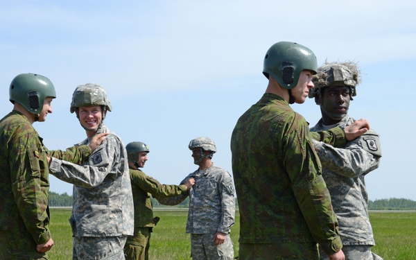 A group of military personnel from at least two different armed forces are interacting on an open grassy field, likely an airfield or training area. Soldiers wearing US Army ACU (Army Combat Uniform) pattern uniforms are paired with soldiers wearing a different camouflage pattern, possibly Estonian or another NATO ally uniform. The interactions appear friendly and cooperative, suggesting a joint training exercise or multinational military event.