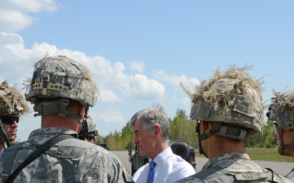 A civilian man in a white dress shirt and blue tie is engaged in conversation with a group of soldiers wearing US Army ACU (Army Combat Uniform) digital camouflage and helmets with ghillie-style camouflage covers. The setting appears to be an outdoor military training area or airfield with trees and a paved surface visible in the background. The interaction suggests a briefing or inspection by a civilian official or dignitary.