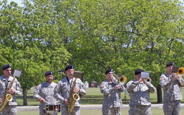 A military band of seven soldiers in US Army combat uniforms (ACUs) and black berets performs outdoors on a grass field. The ensemble includes brass and woodwind instruments along with percussion, suggesting a formal military ceremony or parade. The setting appears to be a military installation with trees and buildings visible in the background.
