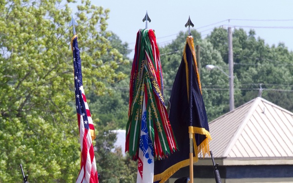 A U.S. Army color guard unit stands in formation on a grass field, presenting multiple flags including the American flag and what appears to be a unit guidon with numerous campaign streamers. The soldiers are wearing Army Combat Uniforms (ACU) with black berets, and two armed guards flank the formation carrying rifles. The ceremony appears to be taking place at a military installation during daylight hours.
