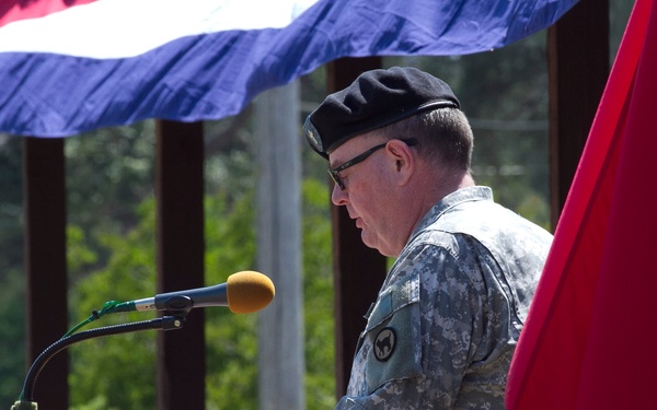 A U.S. Army officer in ACU (Army Combat Uniform) and black beret is speaking at a wooden podium equipped with a microphone during an outdoor military ceremony. A red general officer flag with white stars is visible to his right, and a U.S. flag is partially visible overhead. The setting appears to be a formal military event held outdoors in a wooded area.