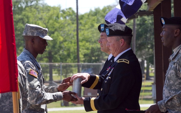 A U.S. Army military ceremony is taking place outdoors, likely a change of command or unit activation/inactivation ceremony. A senior officer in dress blues, identifiable as a Major General by the two-star rank insignia, is handing or receiving a cylindrical case (likely containing a unit guidon or colors) to/from a soldier in ACU (Army Combat Uniform). Additional soldiers in ACUs and black berets are present as part of the formal ceremony.