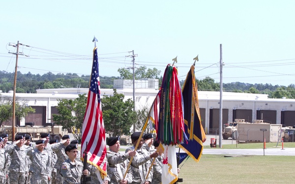 A U.S. Army military ceremony is taking place on a parade field at what appears to be a domestic military installation. Soldiers in ACU (Army Combat Uniform) are standing in formation and rendering salutes while a color guard presents the U.S. flag and multiple unit guidons/colors. A senior officer or official stands to the right, also saluting, facing the formation.