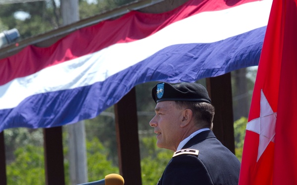 A U.S. Army Major General stands at a wooden podium during an outdoor military ceremony. He is wearing the Army Service Uniform with a black beret bearing a unit flash, and two-star rank insignia is visible on his collar. A red general officer flag with two white stars is displayed to his right, and red, white, and blue bunting decorates the overhead canopy.