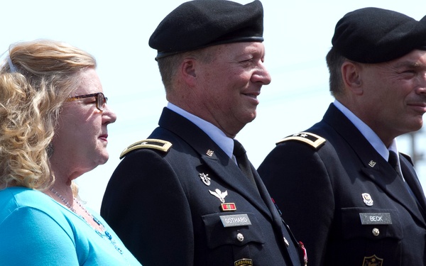 Three individuals stand together outdoors at what appears to be a military ceremony. Two U.S. Army officers in dress uniforms with black berets flank a civilian woman in a turquoise top. Name tags visible on the uniforms read 'GOTHARD' and 'BECK'.