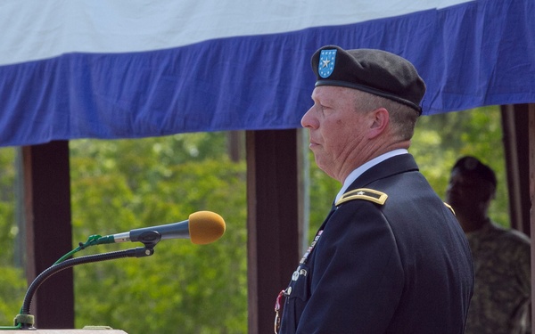 A U.S. Army officer in dress blues uniform stands at a wooden podium delivering a speech at what appears to be a formal military ceremony. He is wearing a black beret with a unit flash insignia and has rank insignia visible on his shoulder. Additional military personnel are visible in the background beneath a blue and white canopy.
