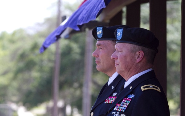 Two U.S. Army officers in dress blue uniforms stand at attention during what appears to be a formal military ceremony. Both wear black berets with a distinctive blue flash bearing a silver star, consistent with XVIII Airborne Corps or a related unit. They are positioned under a covered structure with a blue flag or guidon visible in the background.