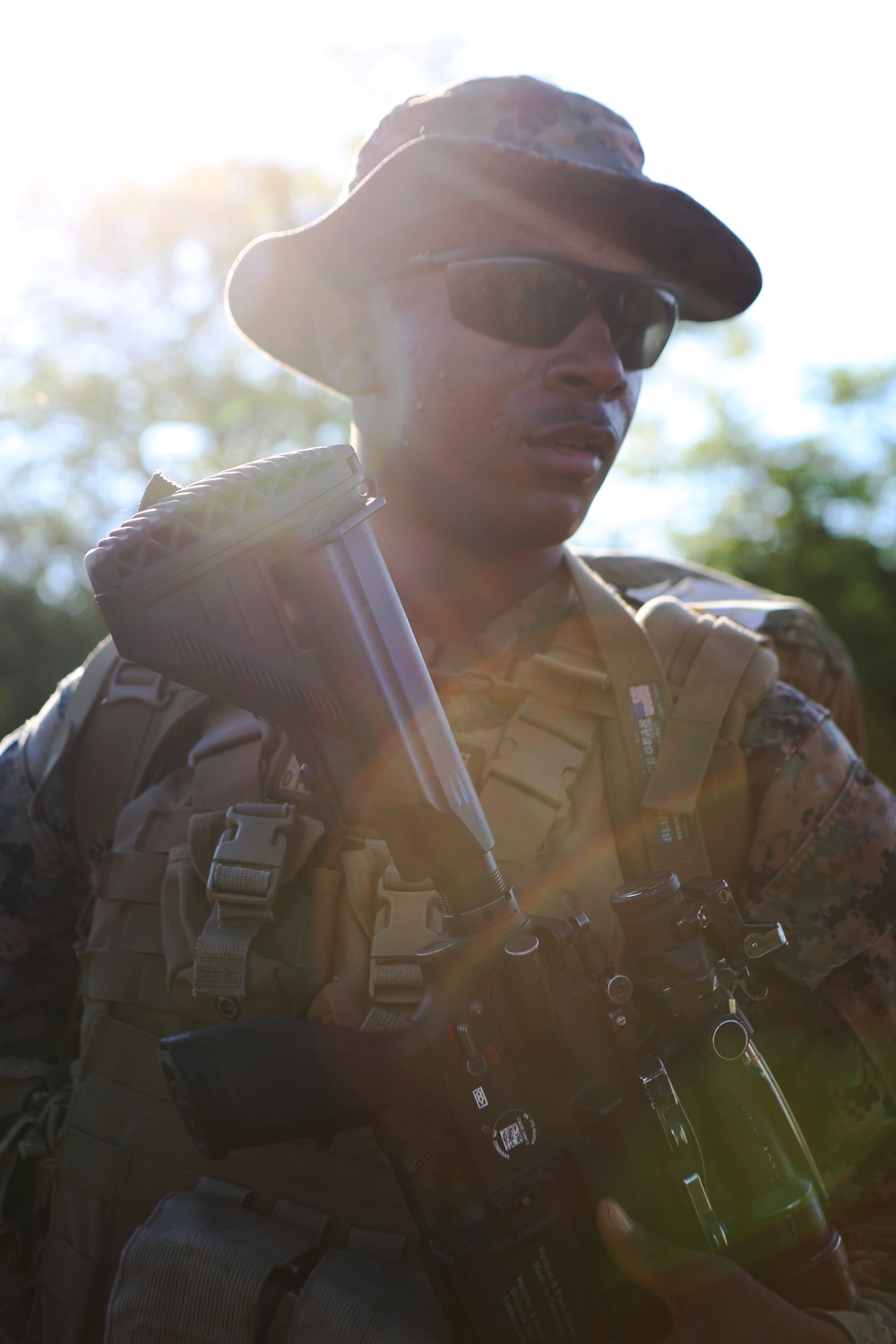 A military service member is photographed in an outdoor setting with strong backlighting creating a lens flare effect. The individual is wearing camouflage uniform and tactical gear while carrying a weapon and what appears to be additional equipment. The dramatic lighting suggests early morning or late afternoon conditions.