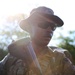 A military service member is photographed in an outdoor setting with strong backlighting creating a lens flare effect. The individual is wearing camouflage uniform and tactical gear while carrying a weapon and what appears to be additional equipment. The dramatic lighting suggests early morning or late afternoon conditions.
