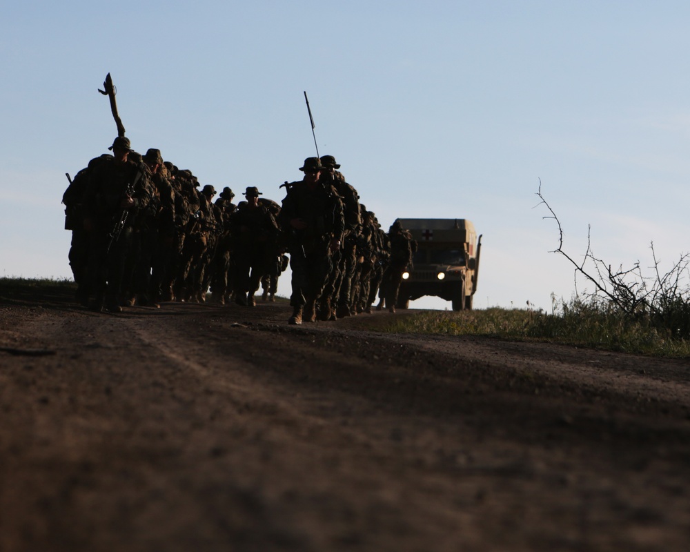 A group of military personnel in full combat gear are conducting a road march along a dirt path, silhouetted against a clear blue sky. A military vehicle, identifiable as a HMMWV (Humvee) with a red cross marking indicating a medical vehicle, follows behind the formation. The low-angle shot and backlighting create strong silhouette effects, suggesting early morning or late afternoon lighting conditions.