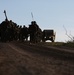 A group of military personnel in full combat gear are conducting a road march along a dirt path, silhouetted against a clear blue sky. A military vehicle, identifiable as a HMMWV (Humvee) with a red cross marking indicating a medical vehicle, follows behind the formation. The low-angle shot and backlighting create strong silhouette effects, suggesting early morning or late afternoon lighting conditions.