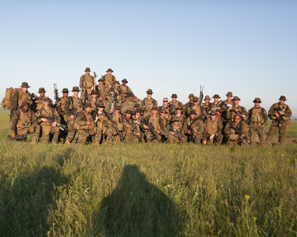 A large group of military personnel, approximately 30-35 individuals, pose for a unit photograph in an open grassy field. All personnel are wearing woodland/desert camouflage uniforms, boonie hats, tactical vests, and are carrying rifles. The photo appears to be taken during golden hour lighting, with the photographer's shadow visible in the foreground.