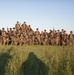 A large group of military personnel, approximately 30-35 individuals, pose for a unit photograph in an open grassy field. All personnel are wearing woodland/desert camouflage uniforms, boonie hats, tactical vests, and are carrying rifles. The photo appears to be taken during golden hour lighting, with the photographer's shadow visible in the foreground.