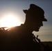 A dramatic silhouette photograph of a person wearing a peaked/flat cap, backlit by a bright setting or rising sun against a blue sky. The subject appears to be holding or standing near a chain, with what looks like a large animal or vehicle partially visible to the left. The strong backlighting creates a high-contrast silhouette effect with visible lens flare.