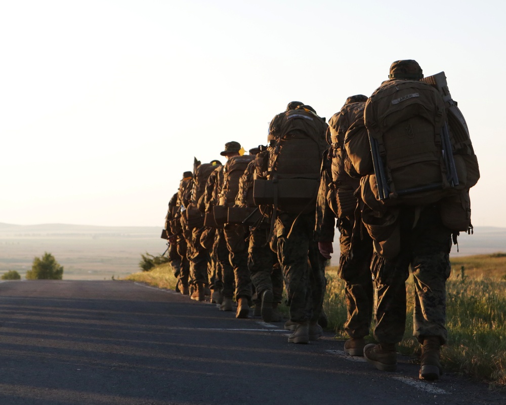 A column of military personnel conducting a road march along a paved road during golden hour (early morning or late evening). The soldiers are fully equipped with large rucksacks, weapons, and combat gear, marching in single file away from the camera. The warm backlighting and open landscape suggest a training exercise or tactical movement in a rural area.