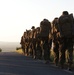 A column of military personnel conducting a road march along a paved road during golden hour (early morning or late evening). The soldiers are fully equipped with large rucksacks, weapons, and combat gear, marching in single file away from the camera. The warm backlighting and open landscape suggest a training exercise or tactical movement in a rural area.