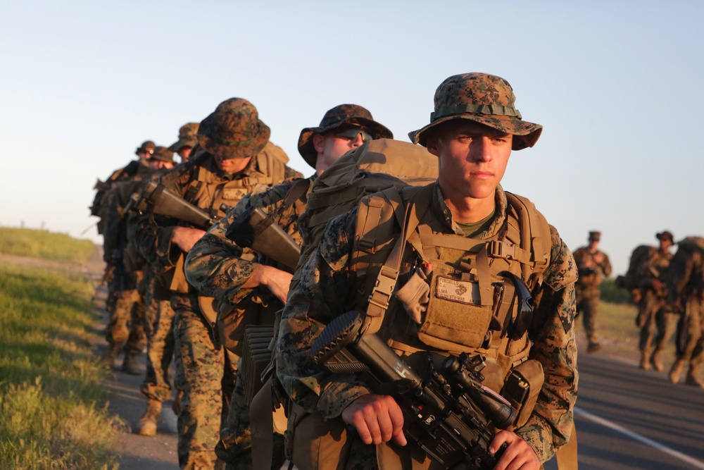 A column of military personnel in full combat gear marches along a paved road during what appears to be a training exercise or tactical movement. The soldiers are wearing MARPAT (Marine Pattern) digital camouflage uniforms and boonie hats, carrying rifles and heavy rucksacks. The warm, low-angle lighting suggests early morning or late afternoon conditions.