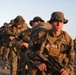 A column of military personnel in full combat gear marches along a paved road during what appears to be a training exercise or tactical movement. The soldiers are wearing MARPAT (Marine Pattern) digital camouflage uniforms and boonie hats, carrying rifles and heavy rucksacks. The warm, low-angle lighting suggests early morning or late afternoon conditions.
