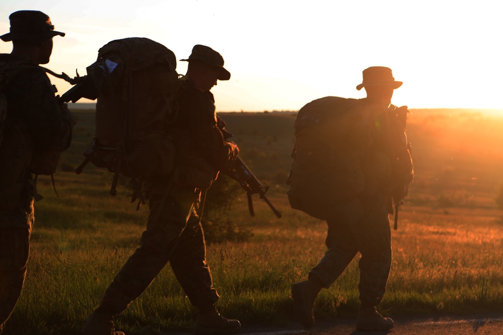 Three silhouetted military personnel are marching across an open field during sunset or sunrise. They are equipped with tactical gear including large backpacks and rifles, moving in a loose formation along what appears to be a dirt path. The strong backlighting creates dramatic silhouettes against a warm orange and yellow sky.