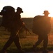 Three silhouetted military personnel are marching across an open field during sunset or sunrise. They are equipped with tactical gear including large backpacks and rifles, moving in a loose formation along what appears to be a dirt path. The strong backlighting creates dramatic silhouettes against a warm orange and yellow sky.