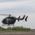 A military medical evacuation (MEDEVAC) helicopter is captured in low hover or landing approach over an open grassy field. The aircraft is painted in olive drab/dark green military livery and displays a red cross symbol, identifying it as a medical transport. The door appears open with at least one crew member visible inside.