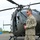 A U.S. Army soldier in ACU (Army Combat Uniform) stands in front of a military helicopter on a tarmac or airfield. The helicopter appears to be a UH-72 Lakota (Eurocopter EC145) marked with a red cross, indicating a medevac/medical evacuation role. The soldier appears to be speaking or briefing someone off-camera.