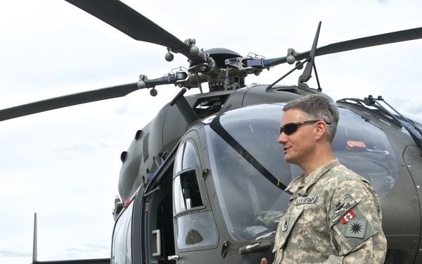 A U.S. Army soldier in ACU (Army Combat Uniform) stands in front of a military helicopter on a tarmac or airfield. The helicopter appears to be a UH-72 Lakota (Eurocopter EC145) marked with a red cross, indicating a medevac/medical evacuation role. The soldier appears to be speaking or briefing someone off-camera.