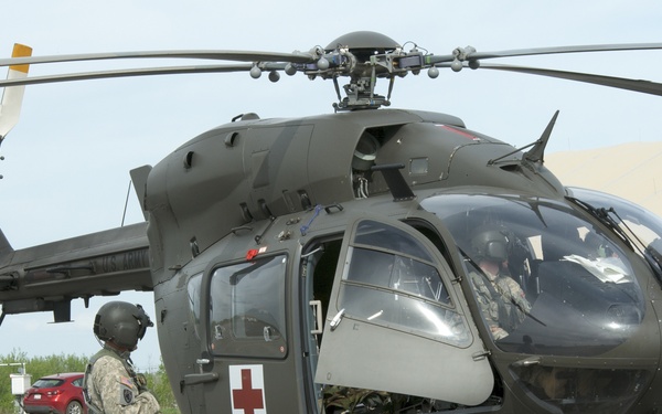 A U.S. Army UH-72 Lakota medical evacuation (MEDEVAC) helicopter is shown on a tarmac or airfield, identifiable by the red cross markings on its fuselage. Several soldiers in Army Combat Uniforms (ACU) are present around the aircraft, with one ground crew member connected via communication cable to the helicopter. The scene appears to be a pre-flight or post-flight ground operation at a military installation.