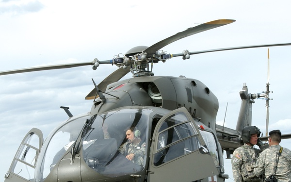 A military medical evacuation (MEDEVAC) helicopter, identifiable by the red cross markings, is on the ground at what appears to be a forward operating base or military airfield. Several soldiers in U.S. Army combat uniforms are gathered around the aircraft, with a pilot visible in the cockpit. The scene suggests a pre-flight briefing, post-landing debrief, or training exercise.