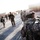 A group of military personnel in U.S. Army Universal Camouflage Pattern (UCP) uniforms are gathered on a paved area, likely a forward operating base or military installation. The foreground soldier, identified by a name tape reading 'LOYA,' is wearing a patrol cap and carrying a large rucksack. The scene appears to depict a troop movement, assembly, or deployment activity in an arid environment.