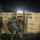 A nighttime military operation showing soldiers in combat gear making a dynamic entry through a doorway in a cinder block wall. The scene is illuminated by artificial lighting, with a bright light source visible in the background sky. At least three soldiers in ACU (Army Combat Uniform) pattern camouflage are visible, moving in tactical formation.