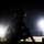 A silhouetted soldier in full combat gear is photographed at night against two bright artificial light sources. The figure is shown in profile, holding what appears to be a rifle, with sandy terrain visible in the background. The dramatic backlighting creates a stark silhouette effect, obscuring most identifying details.