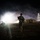 A nighttime military operation scene showing armed soldiers in combat gear moving across an open dirt area. A bright light source illuminates smoke or haze in the background, with low-profile structures and a lattice tower visible. The scene conveys active movement and tactical positioning under low-light conditions.
