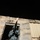 A nighttime photograph showing a figure in military or tactical gear firing a weapon from a doorway of a deteriorated building. The image is motion-blurred, captured at night with muzzle flash visible at the top of the frame. The building appears to be in poor condition with damaged walls and corrugated metal panels.
