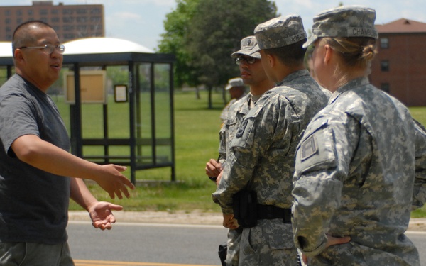 A civilian man in a gray t-shirt is speaking and gesturing with his hands to two U.S. Army soldiers in ACU (Army Combat Uniform) camouflage on what appears to be a military installation. The soldiers are wearing patrol caps and appear to be listening attentively. The setting is outdoors near a road on what looks like a military base, with buildings and trees visible in the background.
