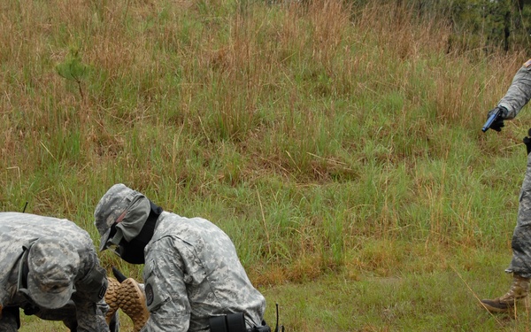 The image shows what appears to be a military training exercise in an outdoor field setting. Three soldiers in US Army ACU (Army Combat Uniform) pattern camouflage are engaged in a detainee handling or ground control drill, while a fourth soldier wearing an 'MP' (Military Police) helmet and holding what appears to be a training weapon observes. All personnel are wearing protective gear including face shields or gas masks, indicating a force-on-force training scenario.