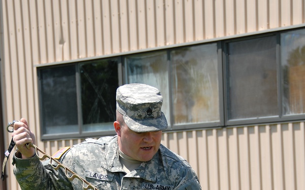 A U.S. Army soldier identified by name tape as 'LANDIS' is kneeling on grass and demonstrating or applying a tourniquet to another soldier who is lying on the ground. The standing soldier is wearing ACU (Army Combat Uniform) with a Staff Sergeant rank insignia and a unit patch on the left shoulder. This appears to be a combat first aid or Tactical Combat Casualty Care (TCCC) training exercise.