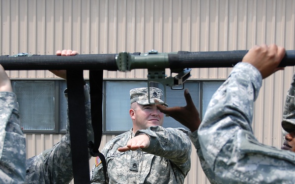 Several U.S. Army soldiers in ACU (Army Combat Uniform) camouflage are handling a large piece of military equipment, likely a mortar or similar weapon system mounted on a bar or rail. A central soldier, identified by a name tape reading 'LANE' and wearing a Staff Sergeant or higher rank insignia, appears to be instructing or supervising the others. The activity appears to be a training exercise taking place outdoors near a military building.
