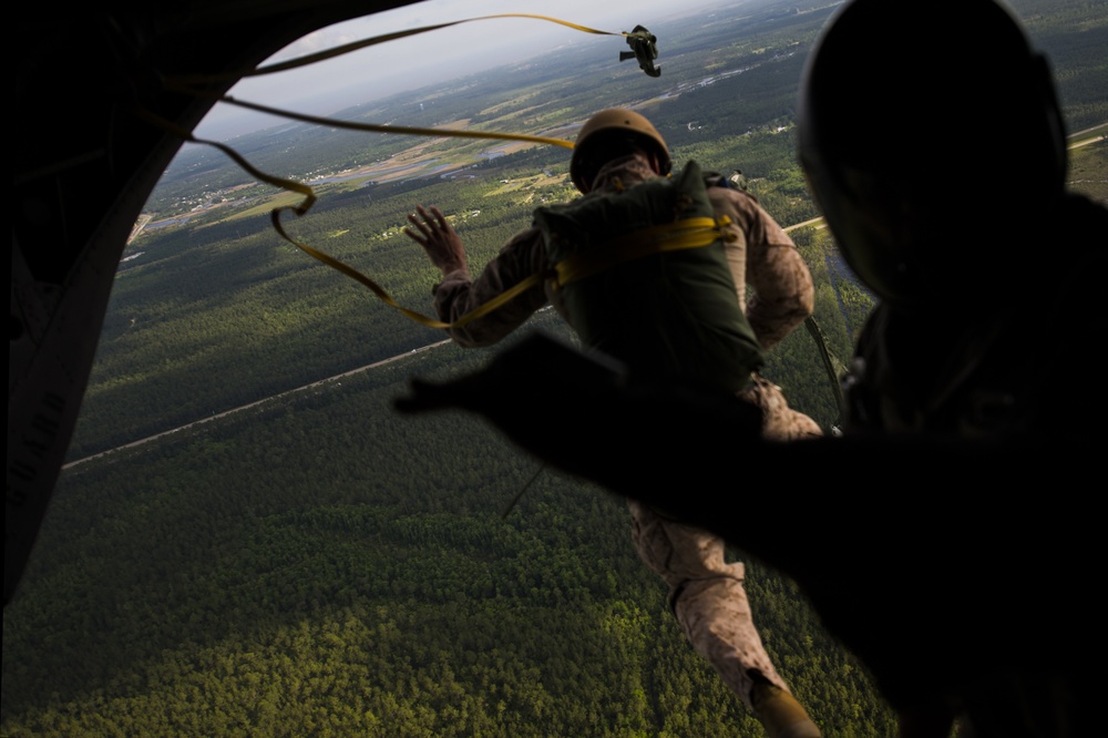 A military paratrooper in camouflage uniform and helmet is exiting an aircraft during a static-line jump exercise, with a yellow static line visible connecting to the aircraft's anchor line cable. A second person in silhouette is visible in the foreground, likely a jumpmaster observing the exit. The terrain below shows dense forested areas with what appears to be an airfield or runway visible in the background.