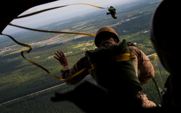 A military paratrooper in camouflage uniform and helmet is exiting an aircraft during a static-line jump exercise, with a yellow static line visible connecting to the aircraft's anchor line cable. A second person in silhouette is visible in the foreground, likely a jumpmaster observing the exit. The terrain below shows dense forested areas with what appears to be an airfield or runway visible in the background.